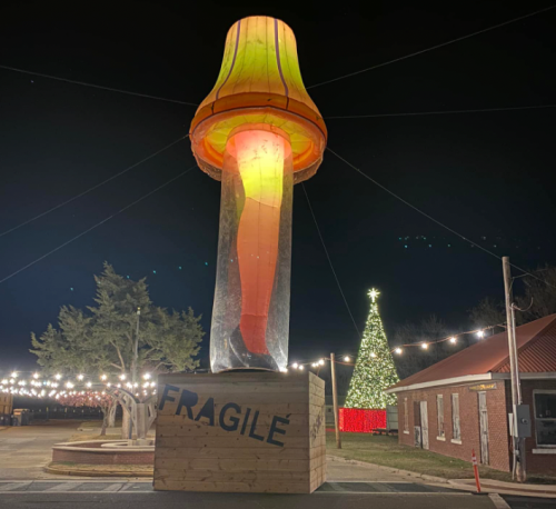 A large, illuminated mushroom sculpture stands at night, with a decorated Christmas tree and string lights in the background.