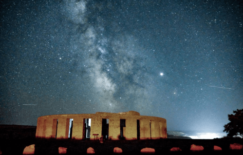 A starry night sky over Stonehenge, with the Milky Way visible and a serene landscape in the background.