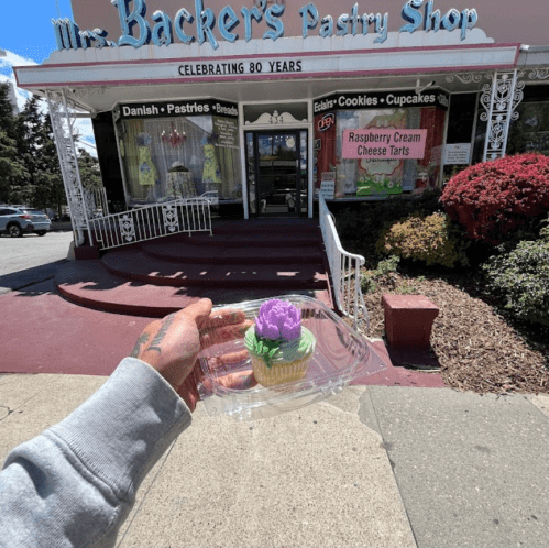A hand holds a cupcake with a purple flower in front of Mrs. Backer's Pastry Shop, celebrating 80 years.