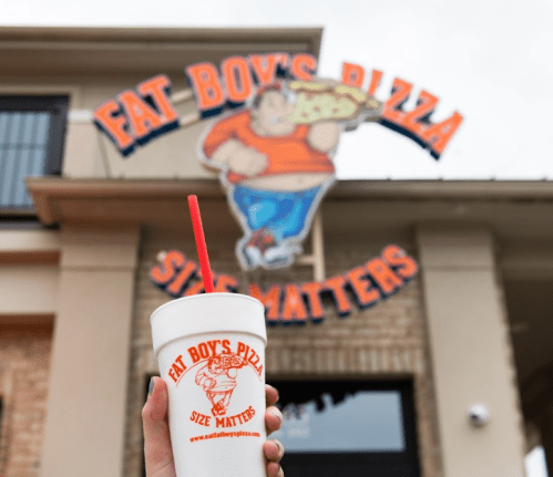 A hand holds a drink cup in front of a pizza shop sign that reads "Fat Boy's Pizza - Size Matters."