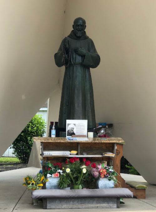 A bronze statue of a monk stands on a pedestal adorned with flowers and a framed photo, under a sheltering structure.