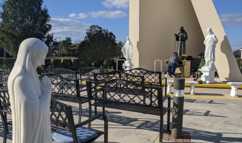 A serene outdoor space with benches, statues of religious figures, and a person tending to a memorial.