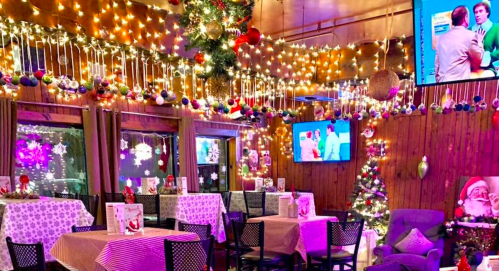 Cozy restaurant decorated for Christmas with lights, ornaments, and a festive tree, featuring tables and TVs.