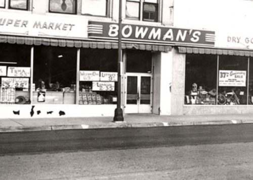 Black and white photo of Bowman's Super Market storefront with signs and displays, featuring a sidewalk and street view.