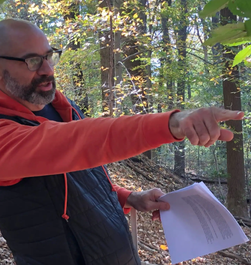 A man in an orange hoodie gestures while holding papers, surrounded by trees with autumn leaves.
