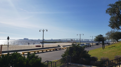 A sunny beach scene with a sandy shore, a bridge in the distance, and a pathway lined with lampposts and greenery.