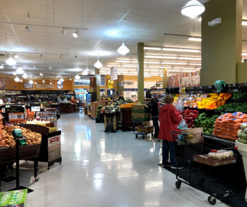 A grocery store aisle filled with fresh produce, shoppers browsing, and colorful fruits and vegetables displayed.