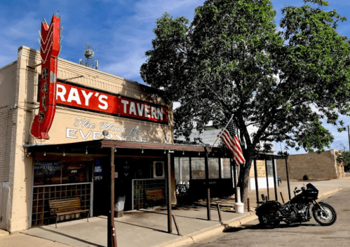 Exterior of Ray's Tavern with a motorcycle parked outside and an American flag, under a clear blue sky.