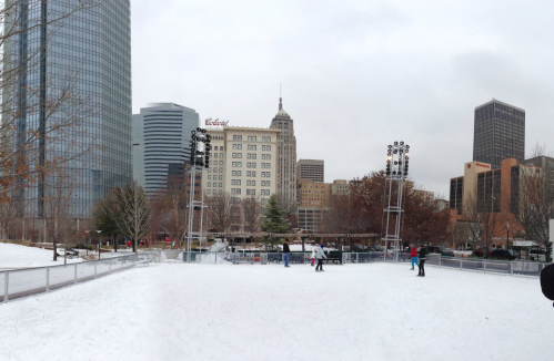 A winter scene of an ice skating rink surrounded by city buildings and trees, with skaters enjoying the activity.