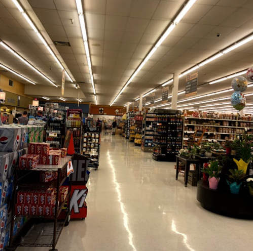 A wide view of a grocery store aisle filled with shelves of products and shoppers, brightly lit with overhead lights.