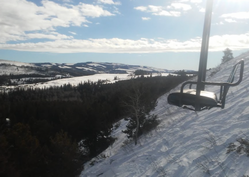 A ski lift chair overlooks a snowy landscape with rolling hills and trees under a partly cloudy sky.