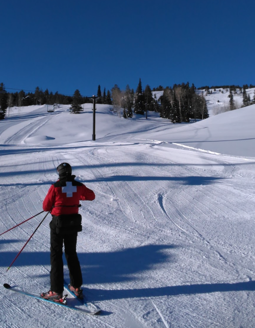 A skier in a red jacket with a white cross stands on a snowy slope under a clear blue sky.