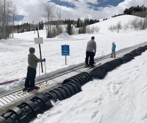 Two children and an adult on skis using a conveyor lift in a snowy landscape, with trees and a blue sky in the background.