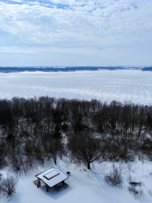 A snowy landscape with a frozen lake and a small shelter surrounded by trees under a cloudy sky.