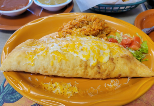 A large cheese-filled burrito topped with melted cheese, served with rice, lettuce, and diced tomatoes on an orange plate.