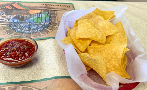 A basket of crispy tortilla chips next to a small bowl of red salsa on a table.