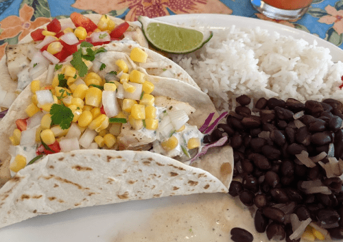 A plate of tacos filled with grilled chicken, corn, and vegetables, served with rice and black beans on the side.