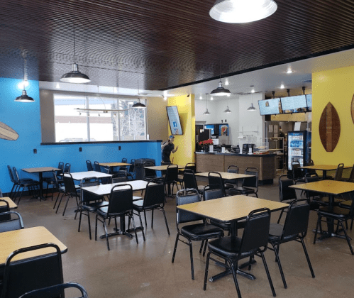 A bright, modern restaurant interior with blue and yellow walls, empty tables, and a counter with digital menus.