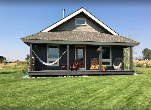 A cozy black house with a porch, hammock, and green lawn under a clear blue sky.