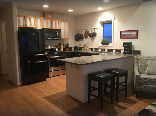 Modern kitchen with black appliances, wooden floors, and a granite countertop with bar stools. Bright and inviting space.