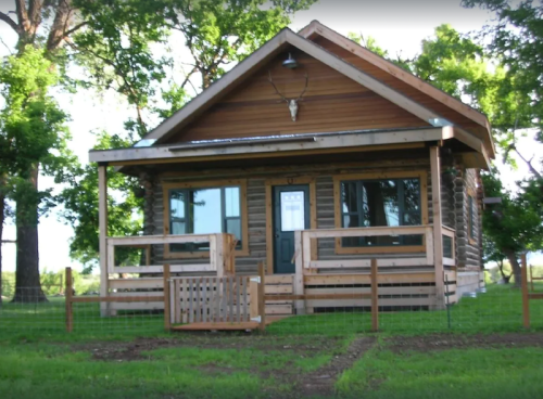 A cozy wooden cabin with a porch, surrounded by trees and a fenced yard.