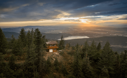 A cozy cabin on a hillside surrounded by trees, overlooking a valley at sunset with a lake in the distance.
