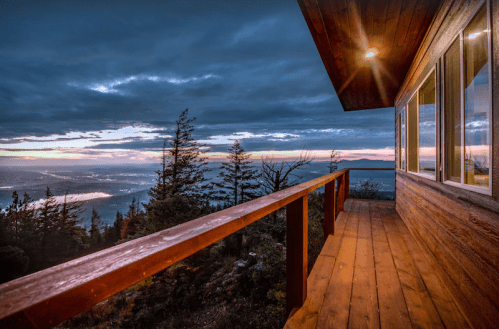A wooden balcony overlooks a scenic view of mountains and valleys at sunset, with dramatic clouds in the sky.
