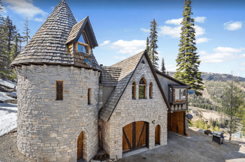 A charming stone house with a turret, surrounded by trees and mountains under a clear blue sky.