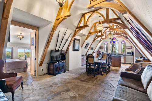 Spacious attic room with wooden beams, stone floor, dining area, and large windows showcasing a scenic view.