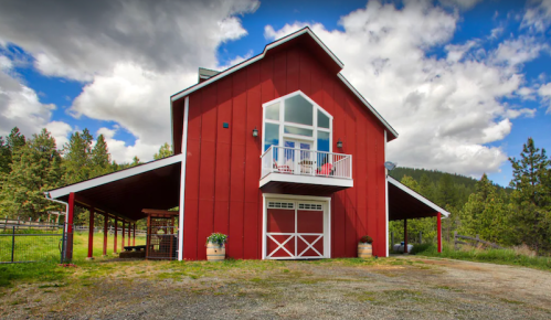 A red barn with a balcony, surrounded by trees and a cloudy sky, set in a rural landscape.