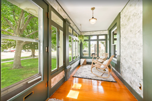 Bright sunroom with large windows, green walls, wooden floor, and two rocking chairs, surrounded by greenery outside.