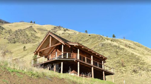 A wooden cabin on a hillside, surrounded by green grass and mountains under a clear blue sky.