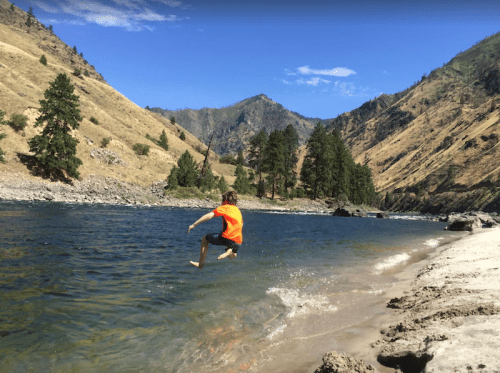 A child in an orange shirt jumps into a river surrounded by mountains and trees on a sunny day.