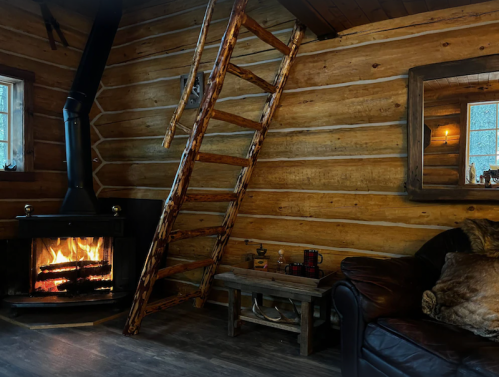 Cozy cabin interior with a wooden ladder, fireplace, and rustic furniture against log walls. Warm and inviting atmosphere.