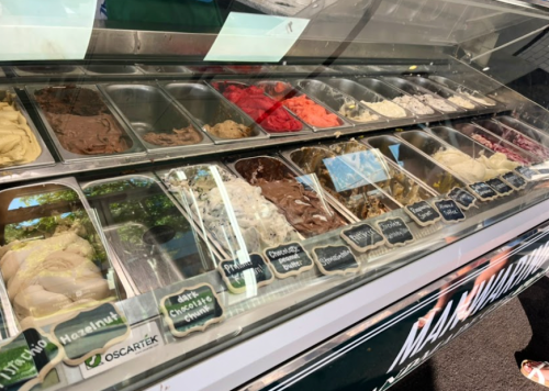 A display of various gelato flavors in metal containers, labeled with names, under a glass case.