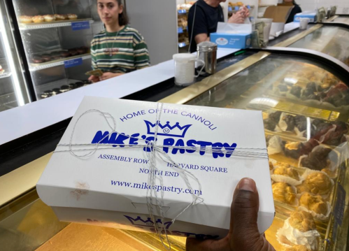 A hand holds a box from Mike's Pastry in front of a display case filled with desserts. A person stands in the background.