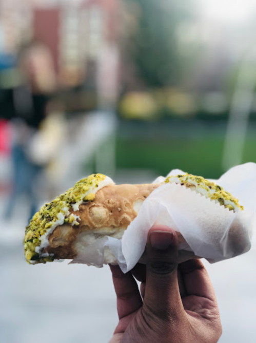 A hand holding a pistachio-topped pastry wrapped in white paper, with a blurred background of greenery.