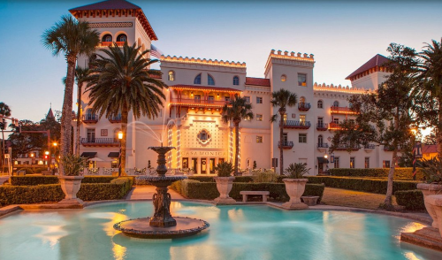 Historic building with a fountain in front, surrounded by palm trees and lush greenery at sunset.