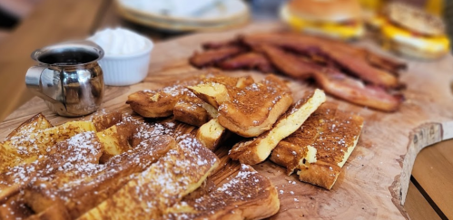 A wooden platter with French toast dusted with powdered sugar, bacon, and a small bowl of whipped cream.