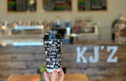 A hand holds a coffee cup with a black and white design in a cozy café setting. Background features a counter and decor.