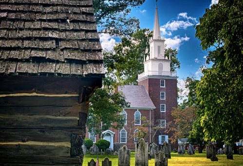 A historic church with a tall steeple stands behind a weathered wooden building and a cemetery under a blue sky.