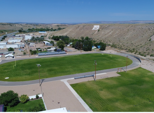 Aerial view of a green sports field surrounded by buildings and hills under a clear blue sky.