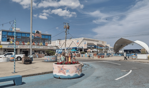 A seaside boardwalk with shops, colorful planters, and people enjoying a sunny day under a blue sky.