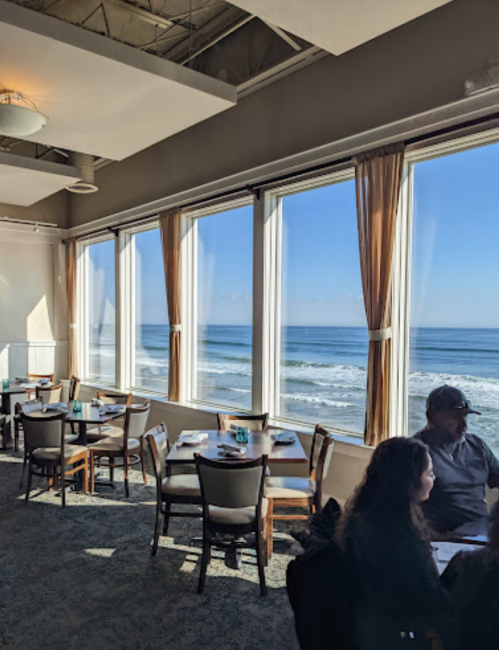 A bright restaurant with large windows overlooking the ocean, featuring tables and chairs, and a couple seated inside.
