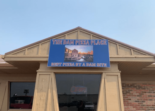 Sign for "The Dam Pizza Place" with the tagline "Best Pizza by a Dam Site" on a building exterior.