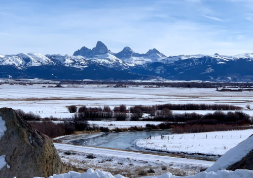 Snow-covered mountains rise in the distance, with a winding river and fields in the foreground under a clear blue sky.
