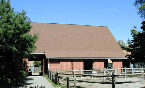 A red barn with a brown roof, surrounded by trees and a wooden fence under a clear blue sky.