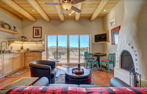 Cozy living space with wooden beams, a fireplace, kitchenette, and a view of the outdoors through large windows.