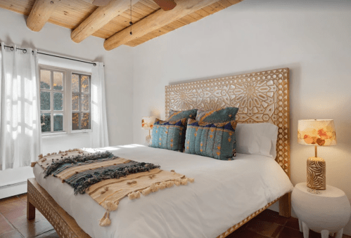 Bright bedroom with a wooden bed, decorative pillows, a cozy blanket, and a lamp, featuring natural light from a window.