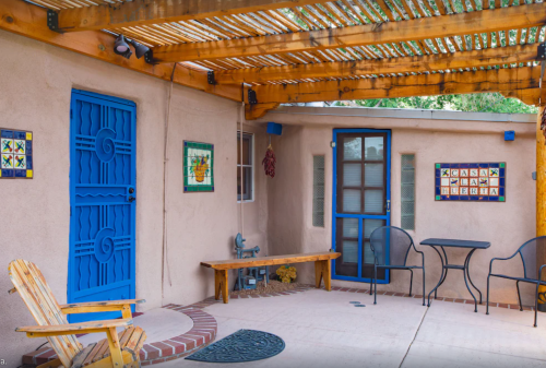 A cozy outdoor patio with a blue door, wooden bench, and two chairs under a wooden pergola. Colorful wall art is visible.